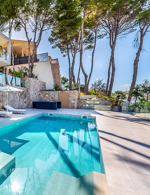 Outdoor swimming pool with white loungers on a paved terrace, set against stone walls and pine trees under a clear blue sky.