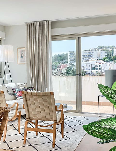 Bright living room featuring rattan armchairs, a patterned area rug, and floor-to-ceiling glass doors opening to a balcony.