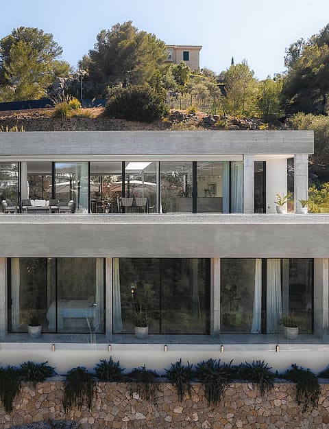 A two-level contemporary villa with exposed concrete, floor-to-ceiling glass, and stone walls on a Mediterranean hillside.