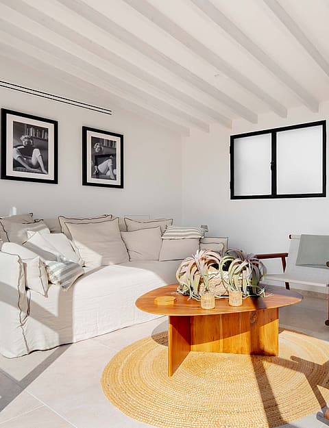 Interior view of a living space with white walls, exposed white ceiling beams, a light-colored sofa, and a wooden coffee table.