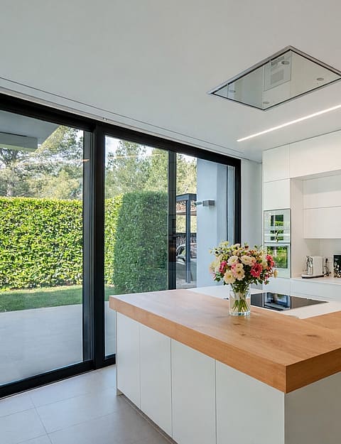 White handleless kitchen with oak-topped island, induction hob and floor-to-ceiling sliding doors to the garden.