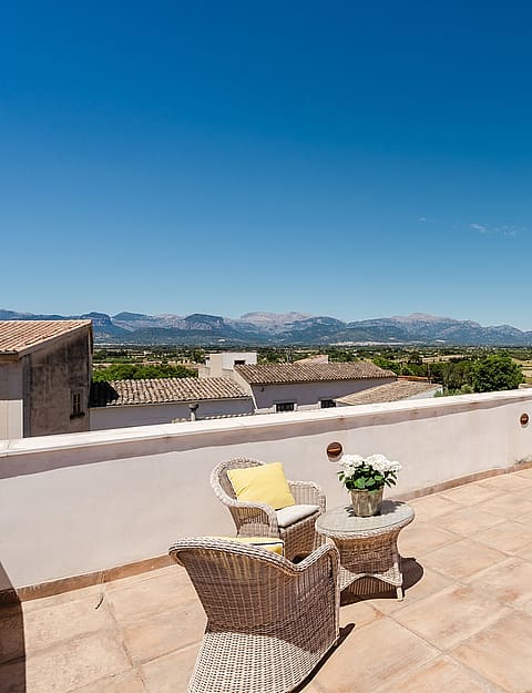 Wide rooftop terrace with terracotta floor tiles, wicker seating, and a dining set, looking out towards distant mountains.