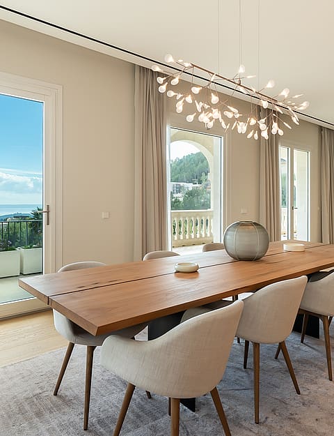 Dining room featuring a long wooden table, light-colored upholstered chairs, and a sculptural chandelier, overlooking the sea.