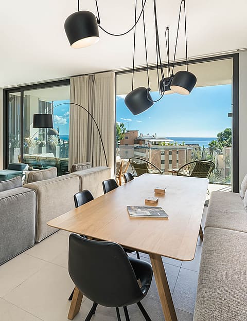 A modern open-plan living room with a large grey sofa, a wooden dining table, and floor-to-ceiling glass doors opening to a terrace with ocean views.