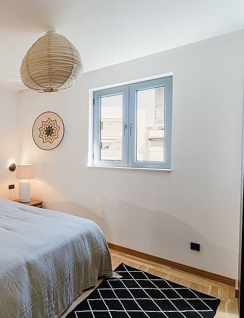 A bright bedroom with a light grey upholstered headboard, beige bedding, a paper pendant lamp, and a geometric patterned rug.