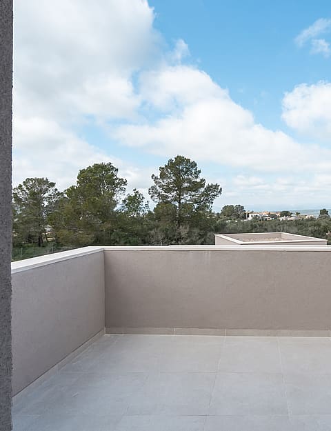 A wide, empty stone-tiled terrace with low perimeter walls, overlooking green trees and distant rolling hills under a blue sky.