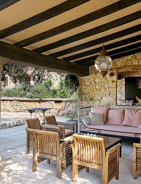 A shaded outdoor patio with exposed timber beams, stone walls, and wooden furniture overlooking a sunlit garden area.