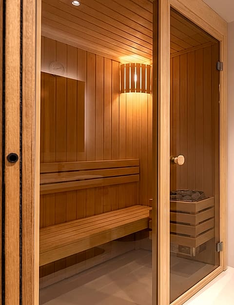 Interior view of a modern sauna featuring light wood paneling, slatted benches, and a glass entry door.