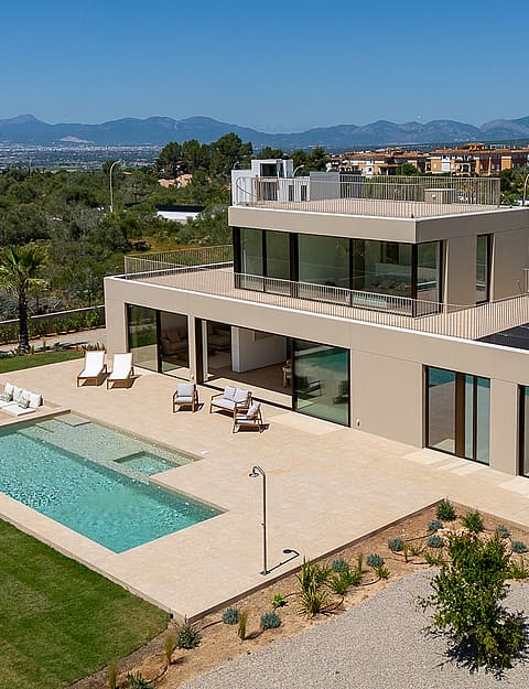 Aerial shot of a beige modern villa with a rectangular swimming pool, terrace seating, and a flat rooftop, set against mountain vistas.