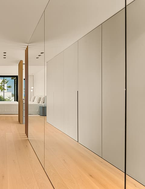 A corridor-style dressing room featuring full-height matte beige wardrobes and a large mirrored wall leading to a bright bedroom.