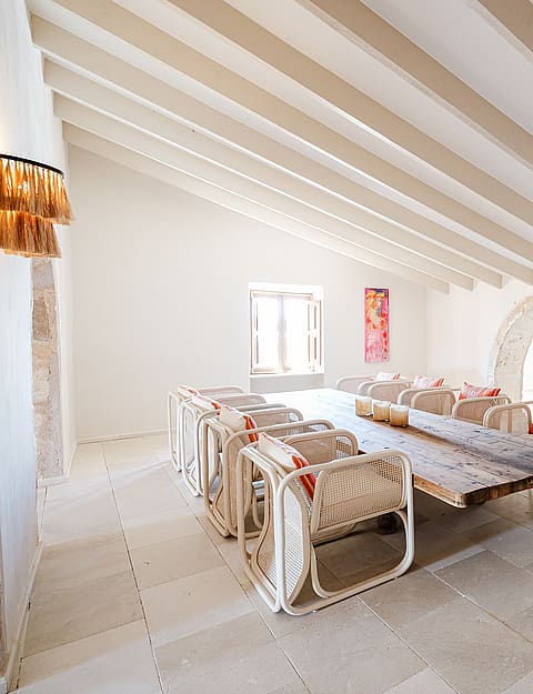 Spacious room with exposed wooden ceiling beams, stone floor, and arched partitions. Long rustic table with woven chairs.