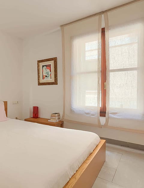 A double bedroom with light wooden bed frame, bedside storage units, and a window with sheer curtains.