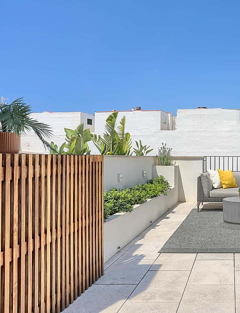 A sunny rooftop terrace featuring light stone flooring, a modular grey sofa, patterned rug, and a wood-slatted privacy screen.