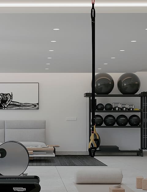 A modern home gym interior featuring a treadmill in the foreground, a suspension trainer, and a bench against a white wall.