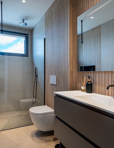 A modern bathroom featuring a floating grey vanity, wood slat wall panels, walk-in shower, and a minimalist white toilet.