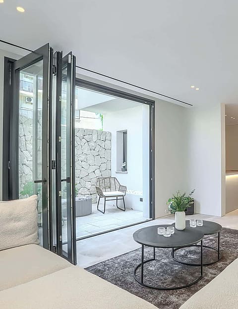 A modern living room with neutral fabric sofas, a coffee table, and folding glass doors leading to a stone-walled terrace.