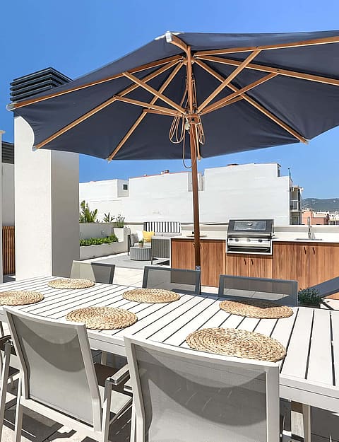 A sunny rooftop terrace in El Molinar featuring a white dining table with woven mats, a large navy umbrella, and built-in grill.