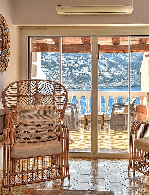 Two rattan armchairs facing sliding glass doors that open onto a sea-view terrace, with decorative mirrors and wall clock.