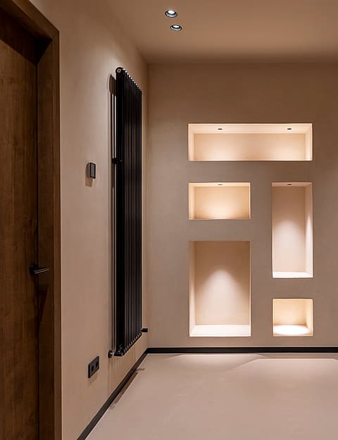 A neutral-toned living space featuring recessed wall shelving with internal warm lighting, a natural wood door, and a low bench.