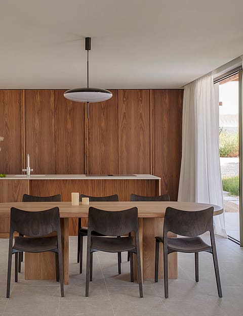 A minimalist dining area featuring an oval wood table and dark chairs, set against full-height walnut kitchen cabinetry.