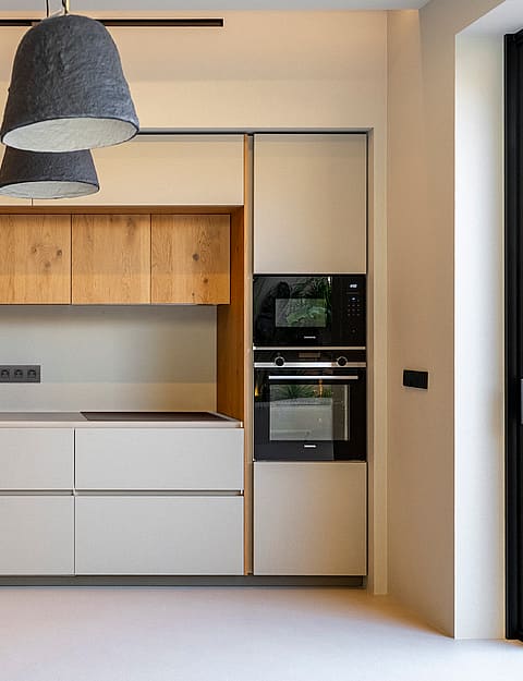 Modern kitchen featuring handleless cream cabinetry, oak upper shelving, integrated appliances, and grey pendant lights.