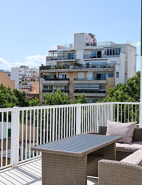 A sunny outdoor terrace with rattan-style sofa seating, white metal railings, and a view of urban residential buildings.