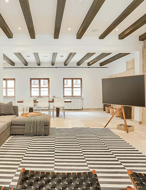 A spacious living room featuring exposed dark wood ceiling beams, white walls, and a large patterned sectional sofa on a striped rug.