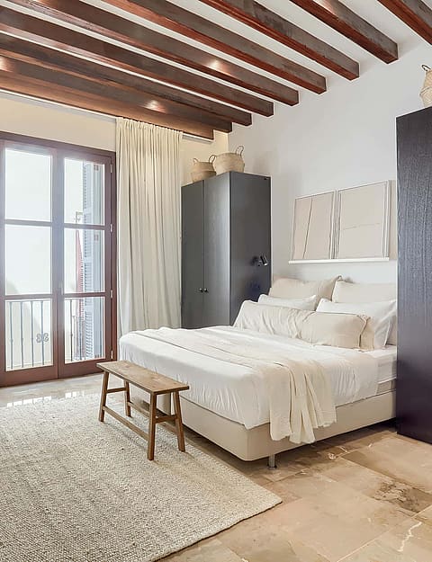 A bright bedroom featuring exposed dark wooden ceiling beams, cream drapes, a beige bed, and dark wood wardrobes.