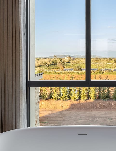 A white freestanding bathtub positioned before a large window looking out onto a rural landscape with distant hills.
