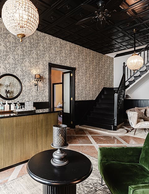 Wide shot of a sophisticated entrance hall featuring a metallic ribbed bar, patterned wallpaper, black ceiling, and green velvet armchairs.