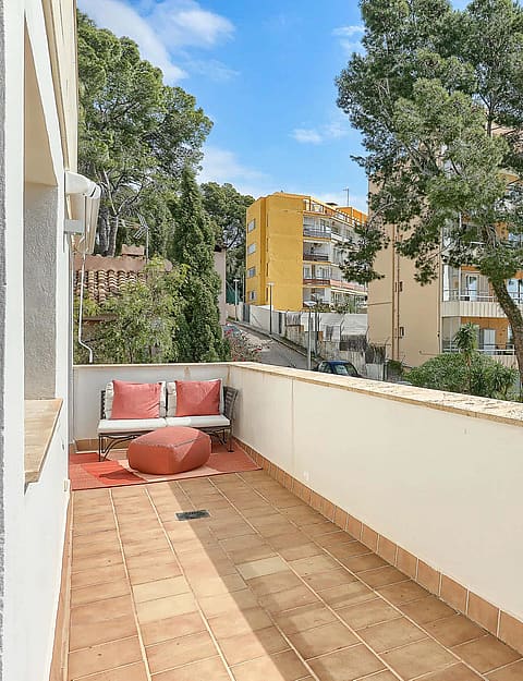 A wide outdoor terrace with terracotta floor tiles, a stone wall, and a small seating area with cushions, facing trees.