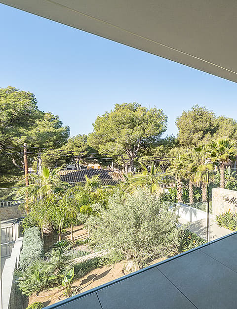 A sheltered terrace with a rough-hewn stone feature wall and glass balustrade, overlooking a Mediterranean garden with pine trees.