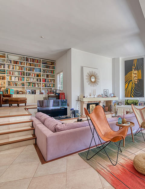 Sunlit living area featuring a split-level layout, large floor-to-ceiling wooden bookshelves, a soft sofa, two leather butterfly chairs, and large glass doors leading to a covered terrace.