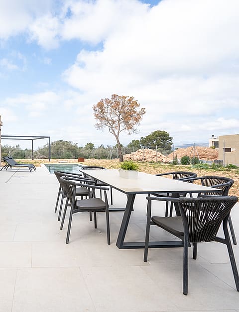 Outdoor dining set on a pale concrete terrace beside a villa wall, overlooking distant mountains under a cloudy sky.