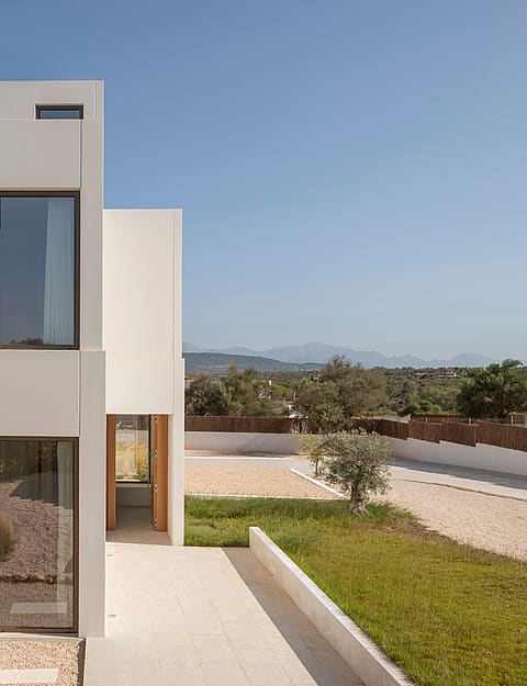 Modern two-story villa featuring white geometric volumes, vertical windows, and wooden shutters under a clear blue sky.