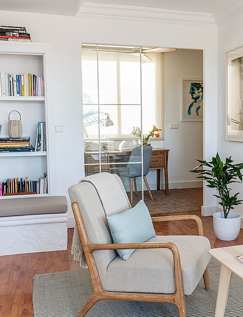 Living room with white walls, wooden floors, built-in bookshelf, and upholstered armchair, looking through a doorway.