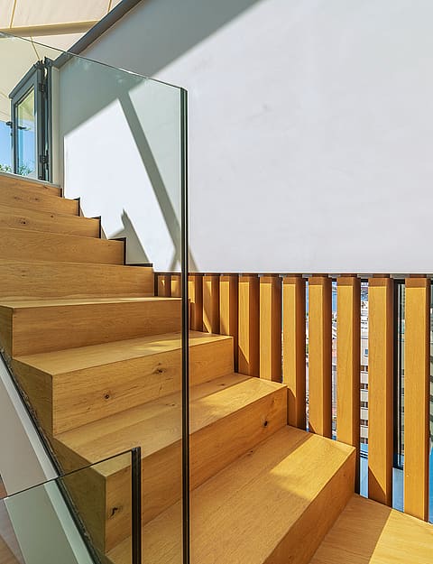 A modern interior staircase featuring solid oak treads and vertical timber slat railings, complemented by a transparent glass balustrade.