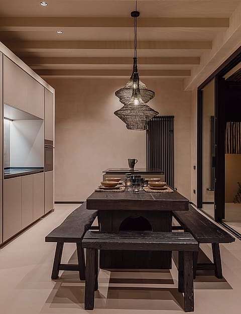 Interior dining space featuring a dark wood table and benches, illuminated by a modern pendant light, opening to an outdoor terrace at night.