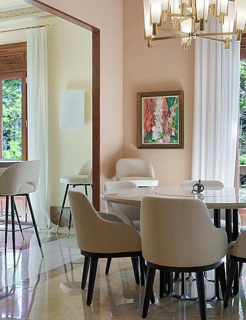 Dining room with a round marble table, upholstered chairs, a crystal chandelier, and large windows overlooking trees.