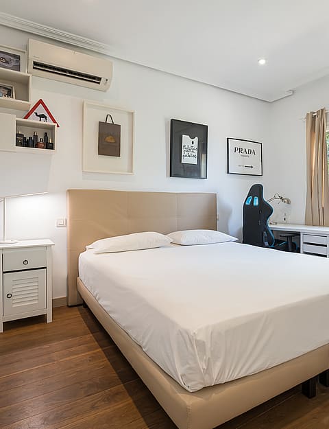 A bright bedroom with a queen-sized bed, white linens, a wooden floor, and a small workspace. A window looks out onto green foliage.