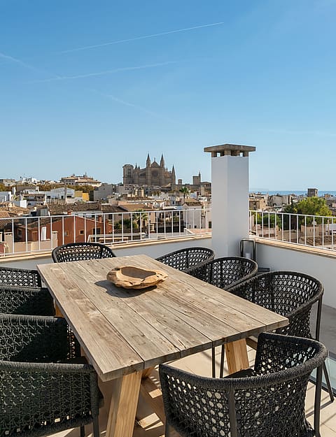 Rooftop terrace with wooden dining table and black chairs overlooking Palma Cathedral and the Mediterranean sea.