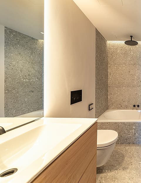 A modern bathroom featuring a white bathtub encased in grey mosaic tiles, a wooden vanity, and a black heated towel rail.