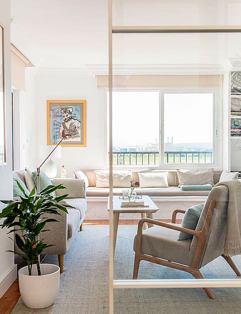 A view through a glass partition into a bright living room with a neutral sofa, patterned area rug, and an armchair.