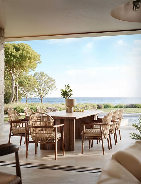 Outdoor dining set with a long wooden table and woven chairs on a covered terrace, overlooking the sea and garden.