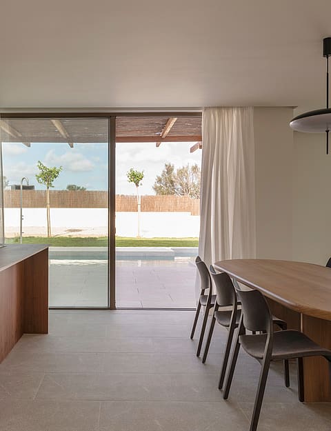 An oval oak dining table and dark chairs in a sunlit room with stone floors and floor-to-ceiling glass sliding doors.