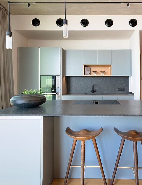 Contemporary kitchen featuring a grey island, dark stone countertops, wooden bar stools, and track lighting in a penthouse.