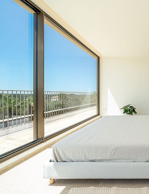 Modern bedroom with a double bed, floor-to-ceiling glass wall, white walls, and view of the exterior landscape.