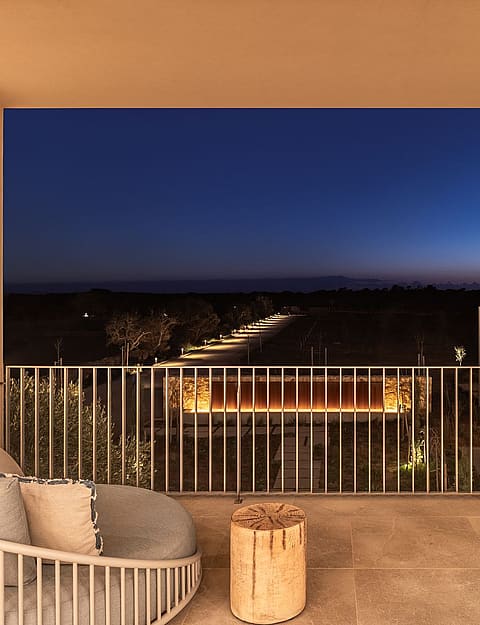 A covered terrace with a lounge chair and wooden side table overlooking a landscape at dusk, featuring a metal railing.