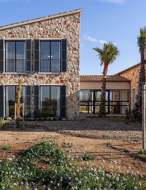 Exterior view of a two-story stone finca with a mono-pitched tile roof and dark-framed windows set in a rural landscape.