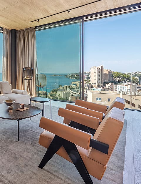 Interior view of a living room featuring modular cream sofa, leather armchairs, and floor-to-ceiling glass walls with sea views.